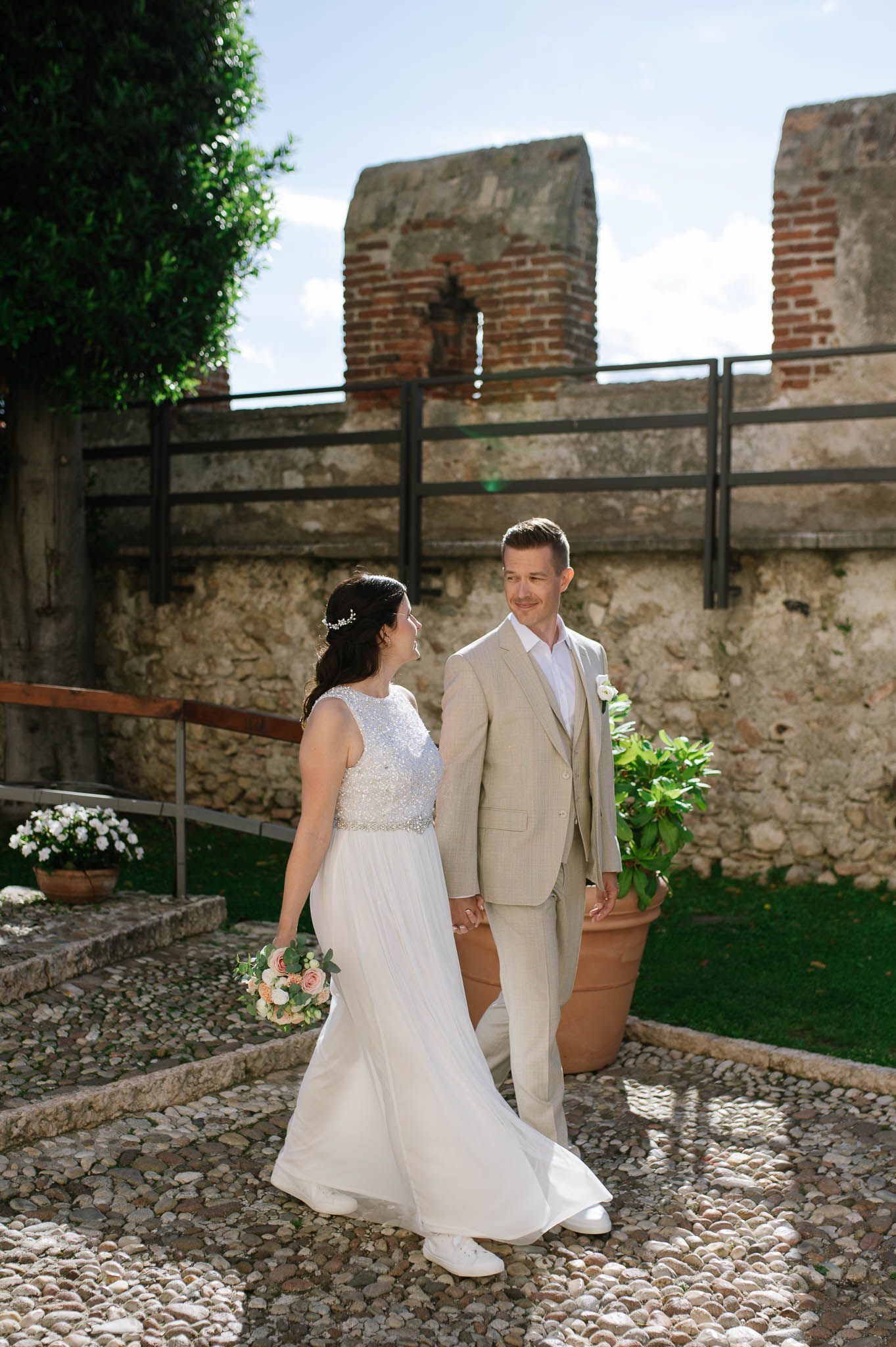 Bride in white lace gown and groom in tan suit walk together at a rustic castle wedding.