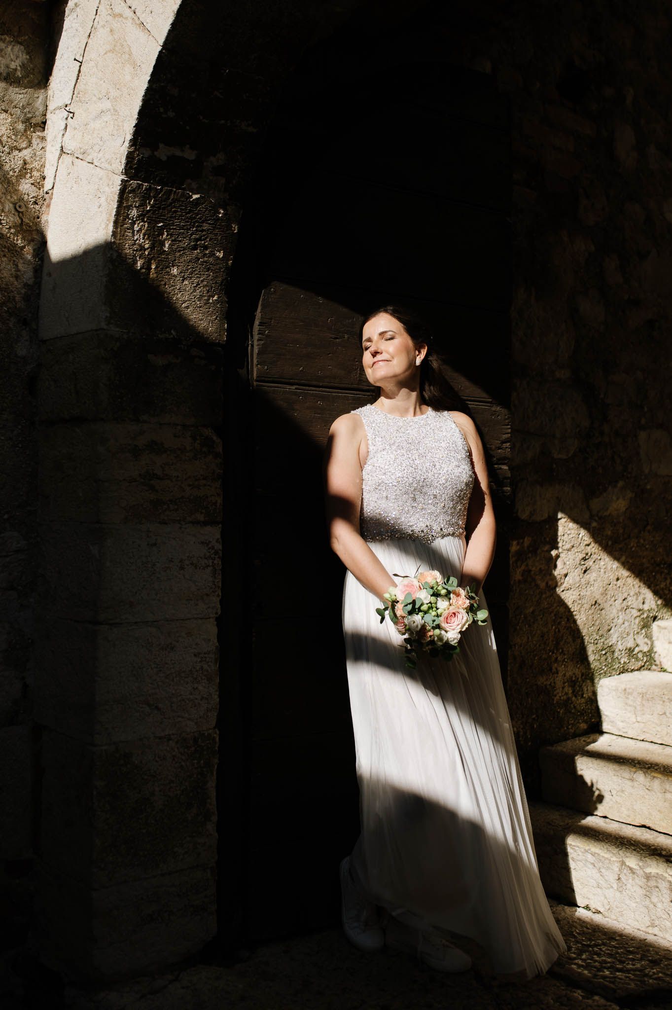 Bride in beaded wedding gown holding pink bouquet standing in dramatic sunlight within ancient stone archway.
