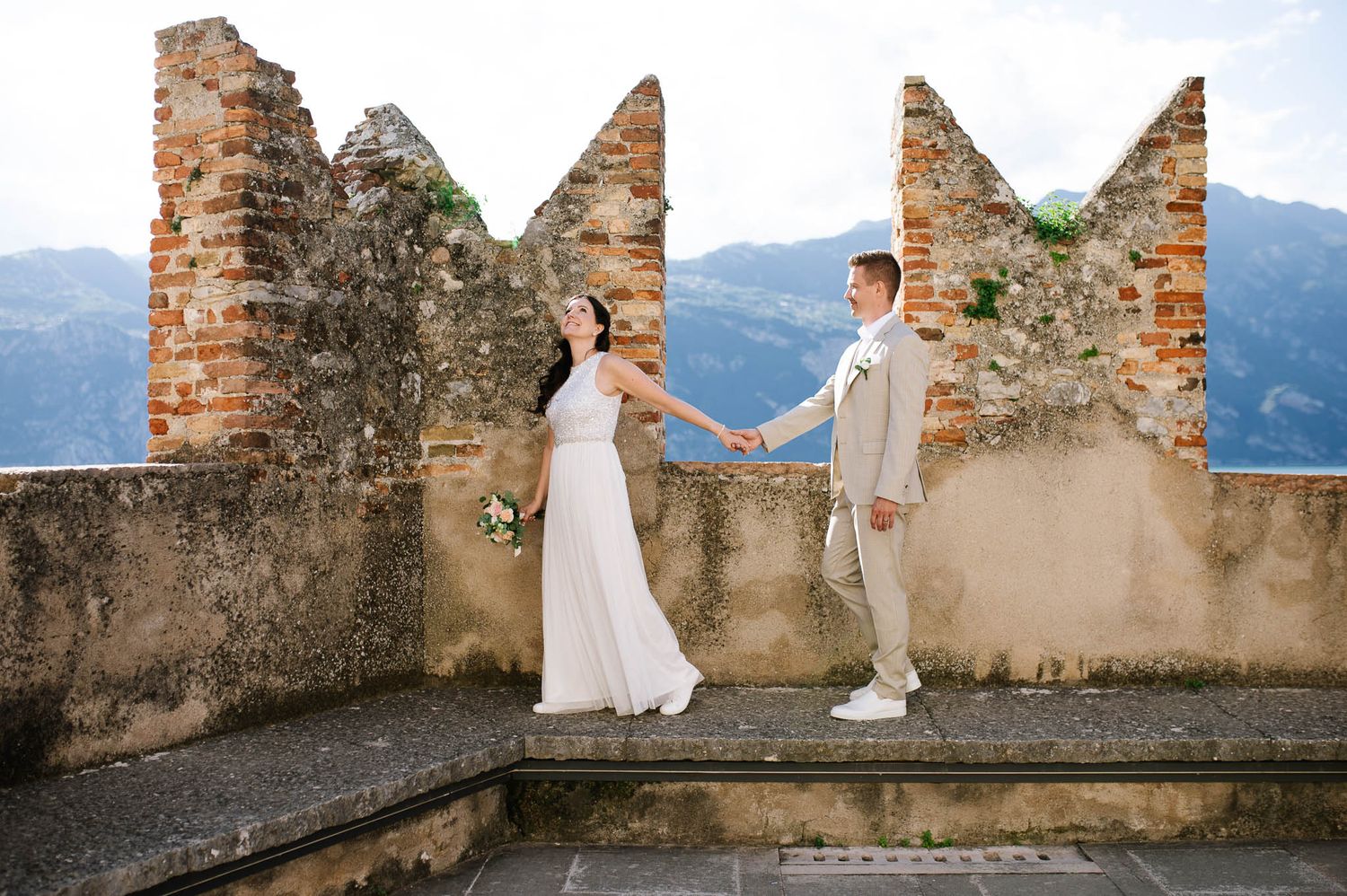 Bride and groom hold hands on a rustic castle terrace with stunning mountain views on their wedding day.