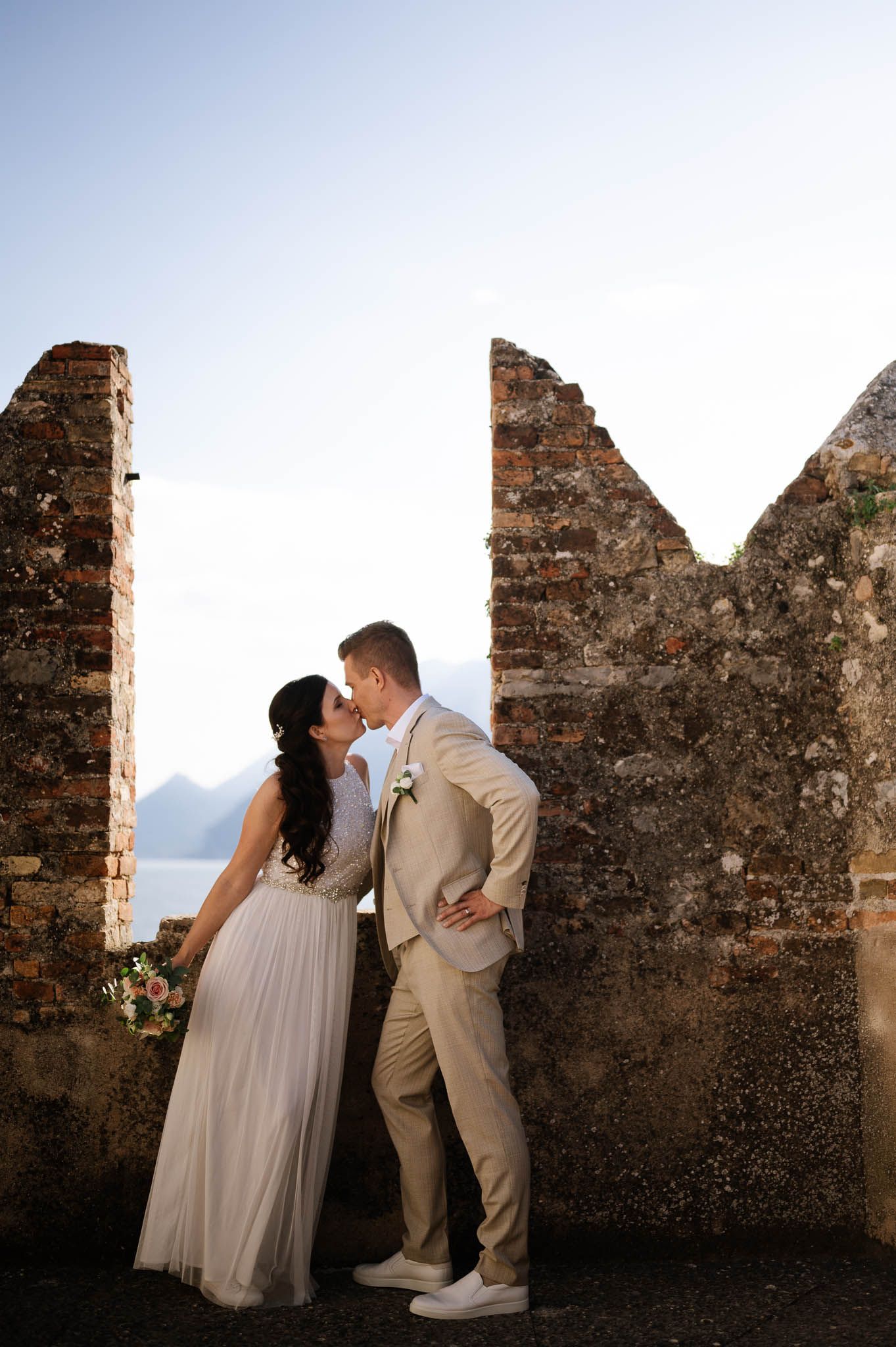 Bride and groom sharing a kiss between ancient stone castle walls with lake views.