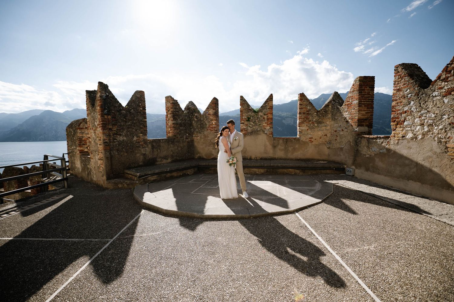 Bride and groom embracing atop a medieval castle terrace with stunning lake and mountain views on their wedding day.