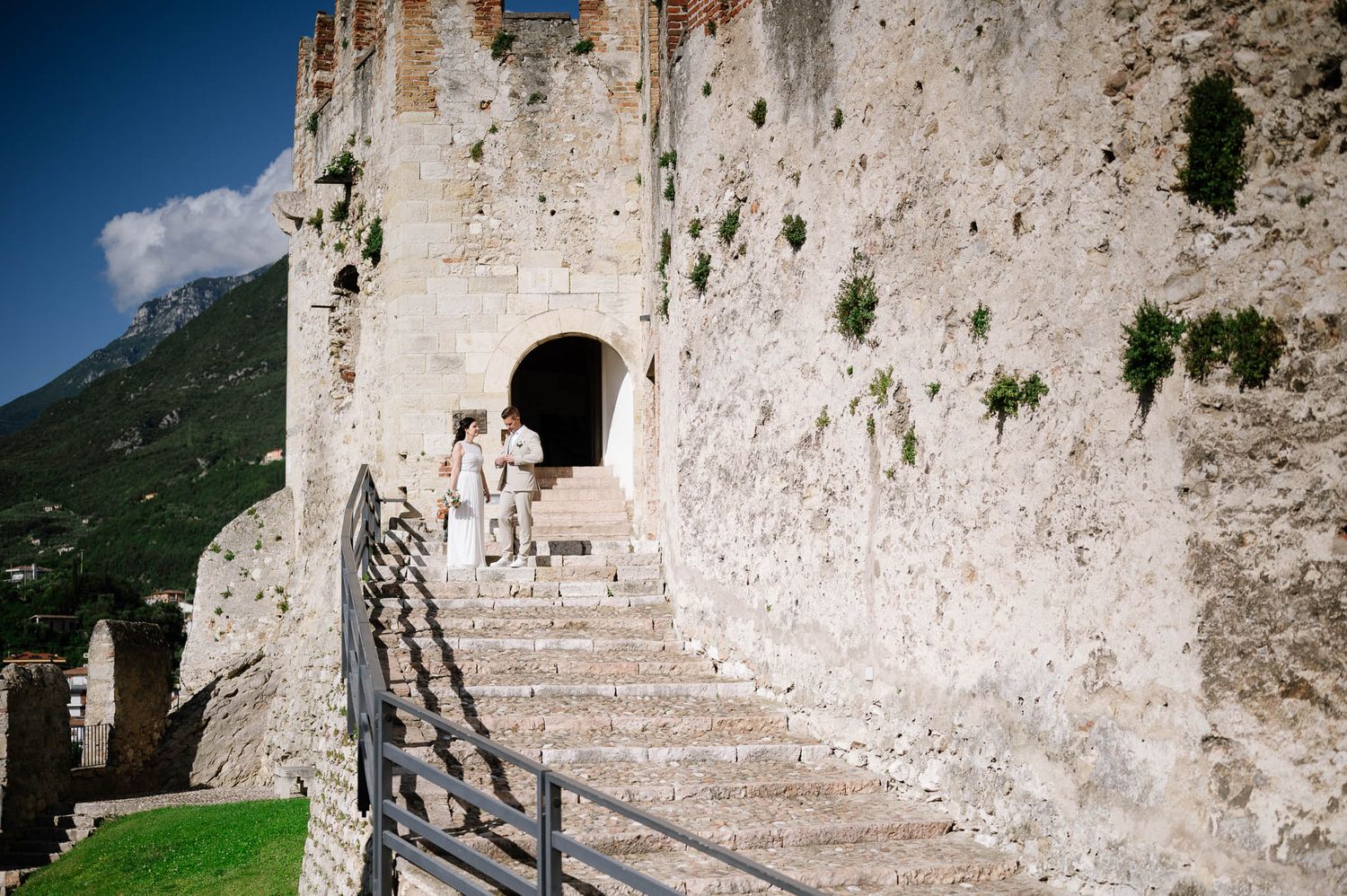 Wedding couple walks through ancient stone castle archway on rustic stairs in scenic Italian landscape.