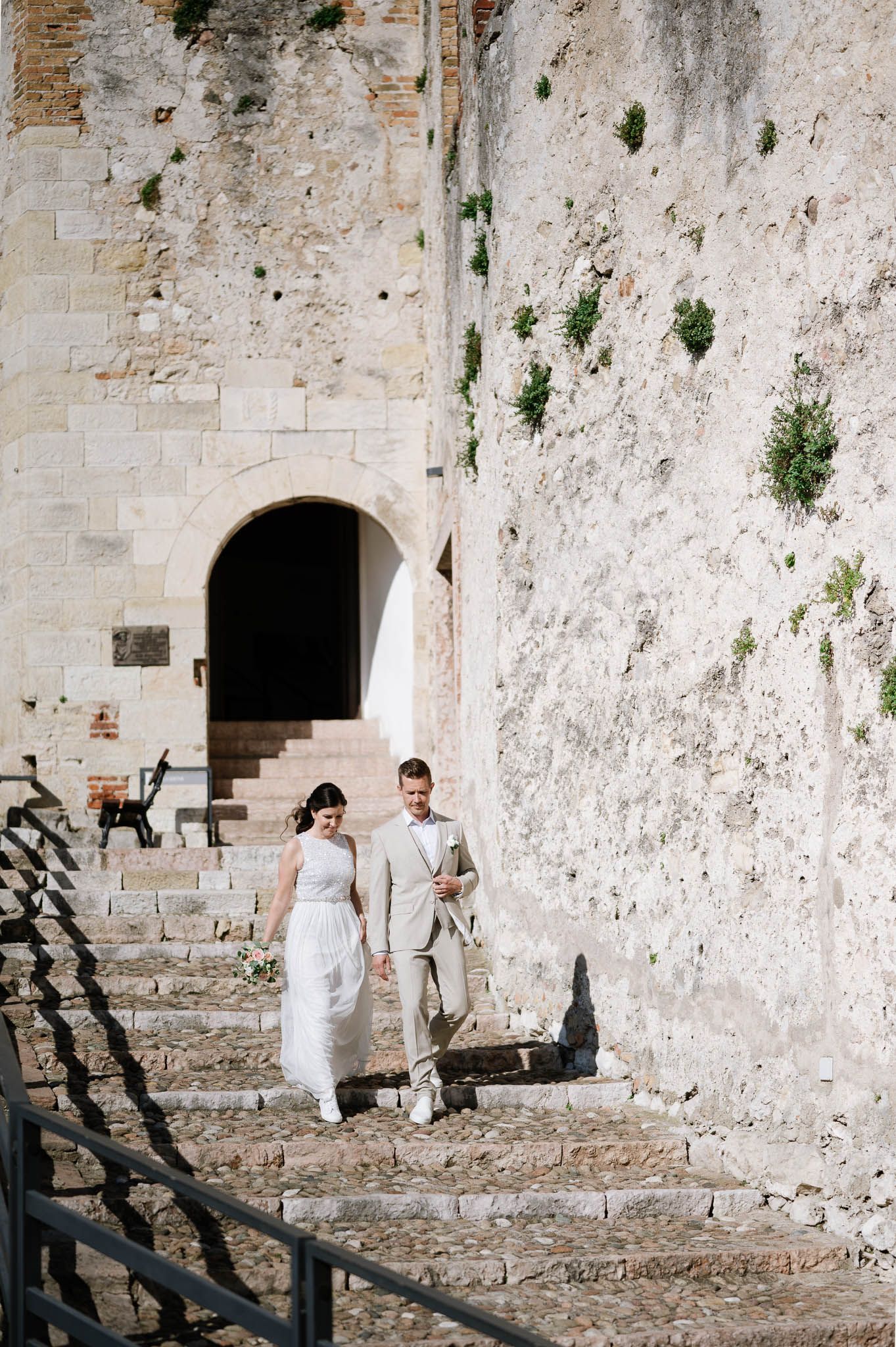 Bride and groom walking down stone steps hand in hand beside rustic historic white stone castle wall.