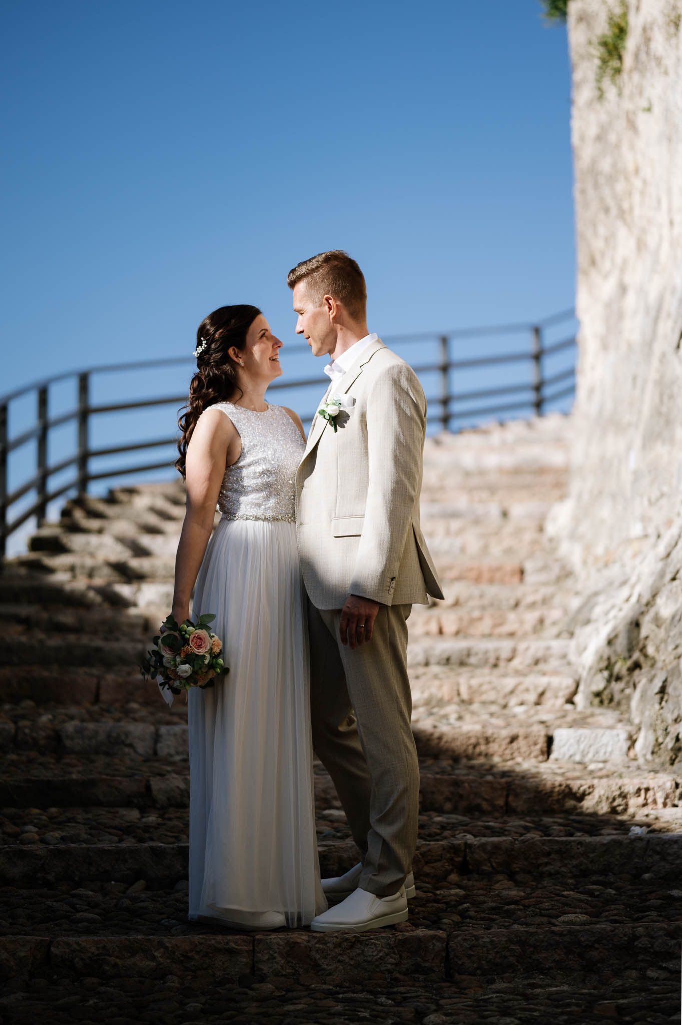 Bride and groom stand together on ancient stone steps under a clear blue sky on wedding day.