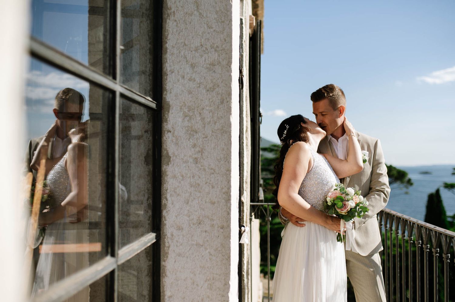 Bride and groom share a tender kiss on a balcony with scenic lake views reflected in window.