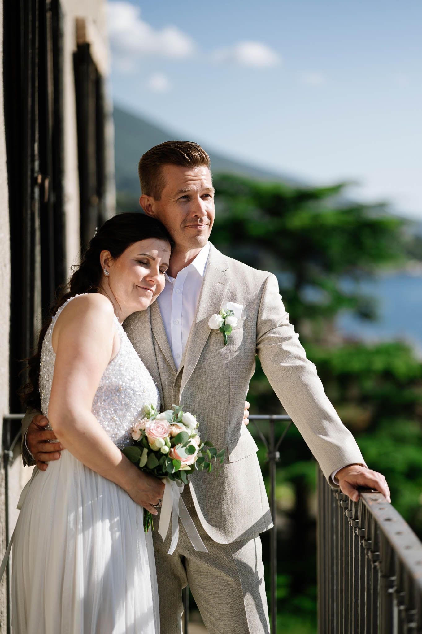 Bride and groom embrace on a balcony overlooking lush greenery and mountains on their wedding day.