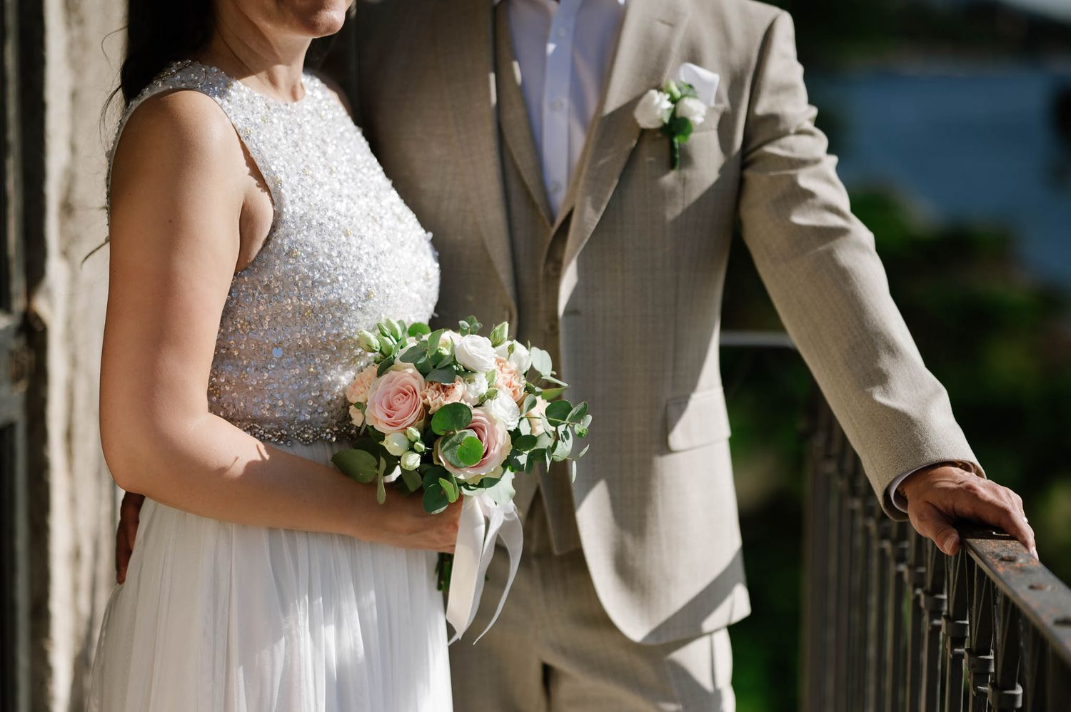 Bride holding pink rose bouquet in beaded gown beside groom in tan suit at railing.