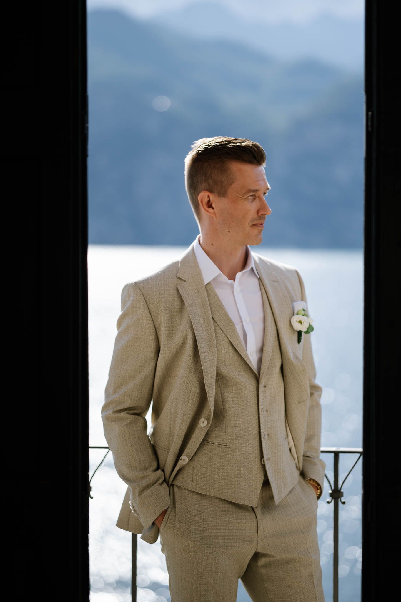 Elegant groom in a beige three-piece suit with boutonniere standing by a balcony overlooking a scenic mountain lake.
