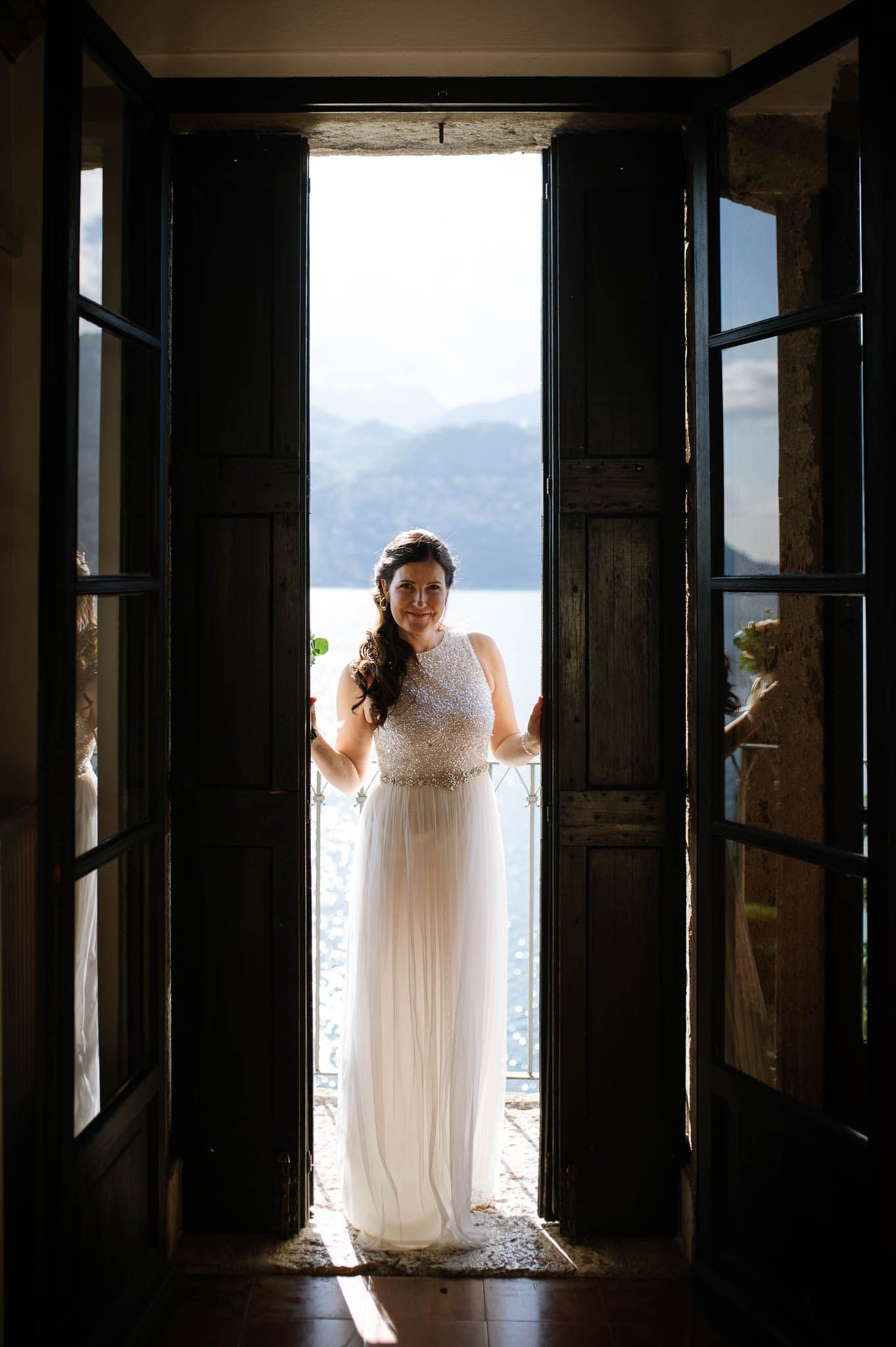Bride in white lace gown standing in open French doors with mountain lake view behind her.