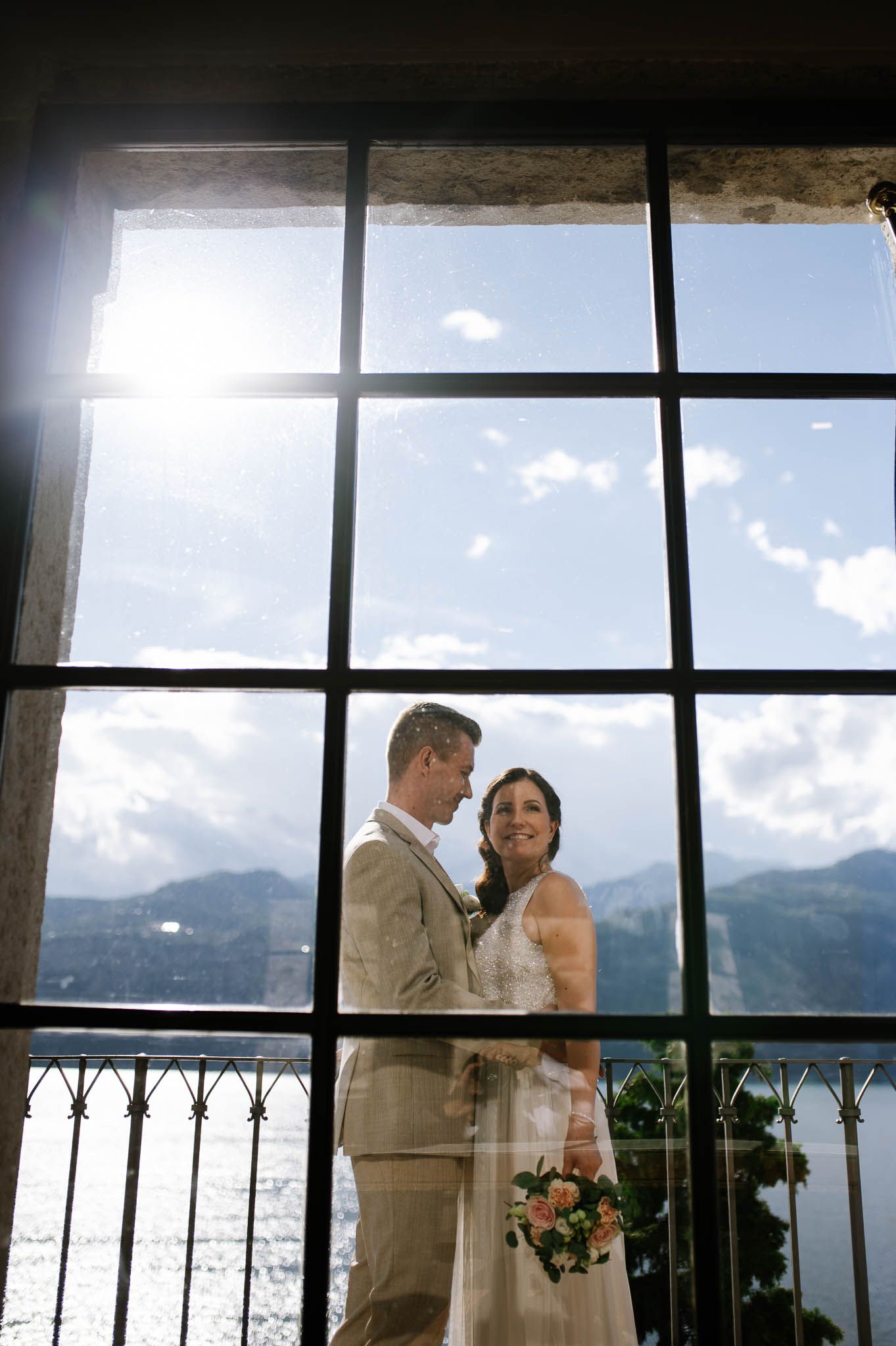 Bride and groom embrace before large window with mountain lake view on their wedding day.