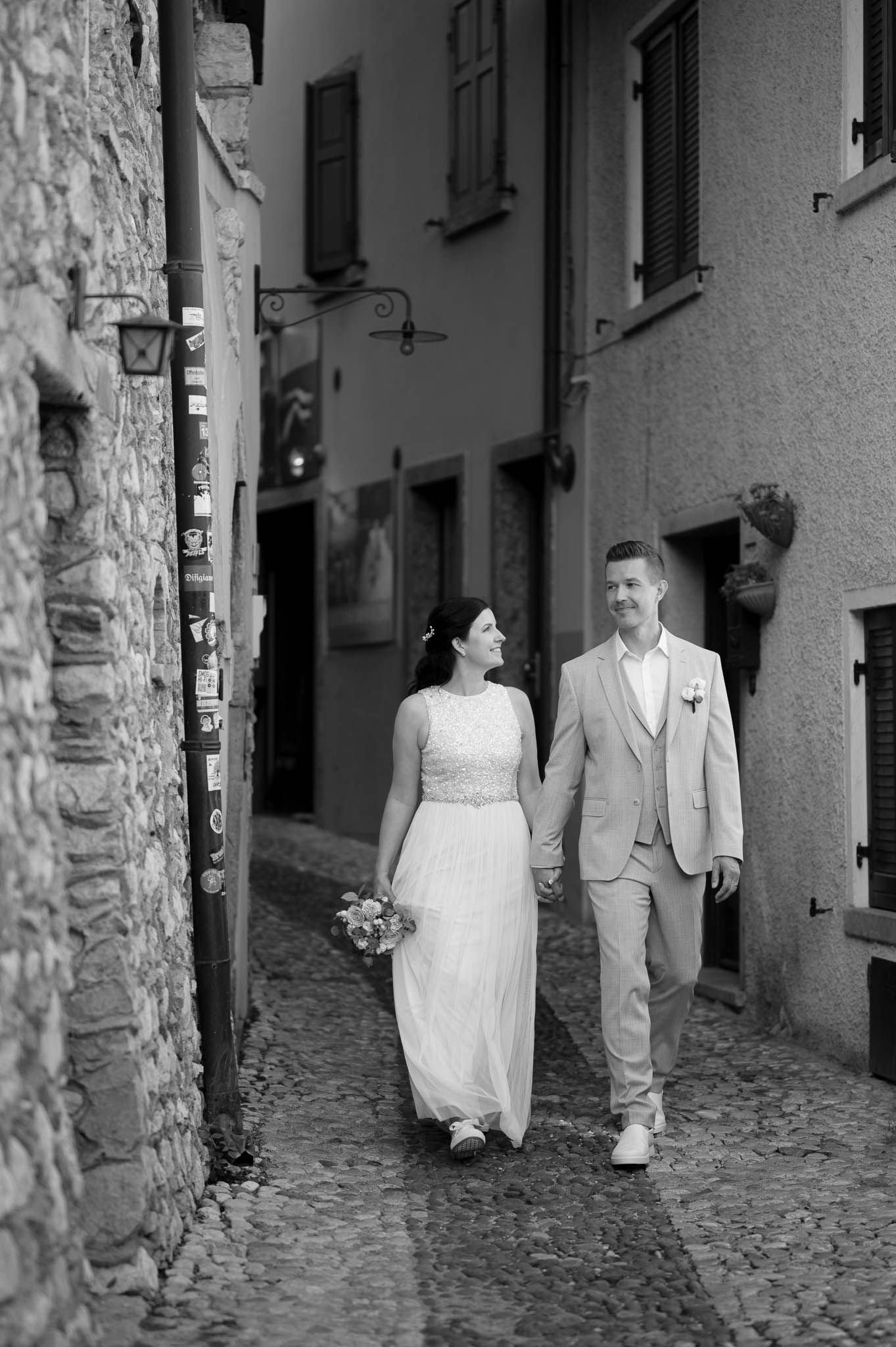 Black and white photo of bride and groom holding hands walking through charming cobblestone Italian village street.