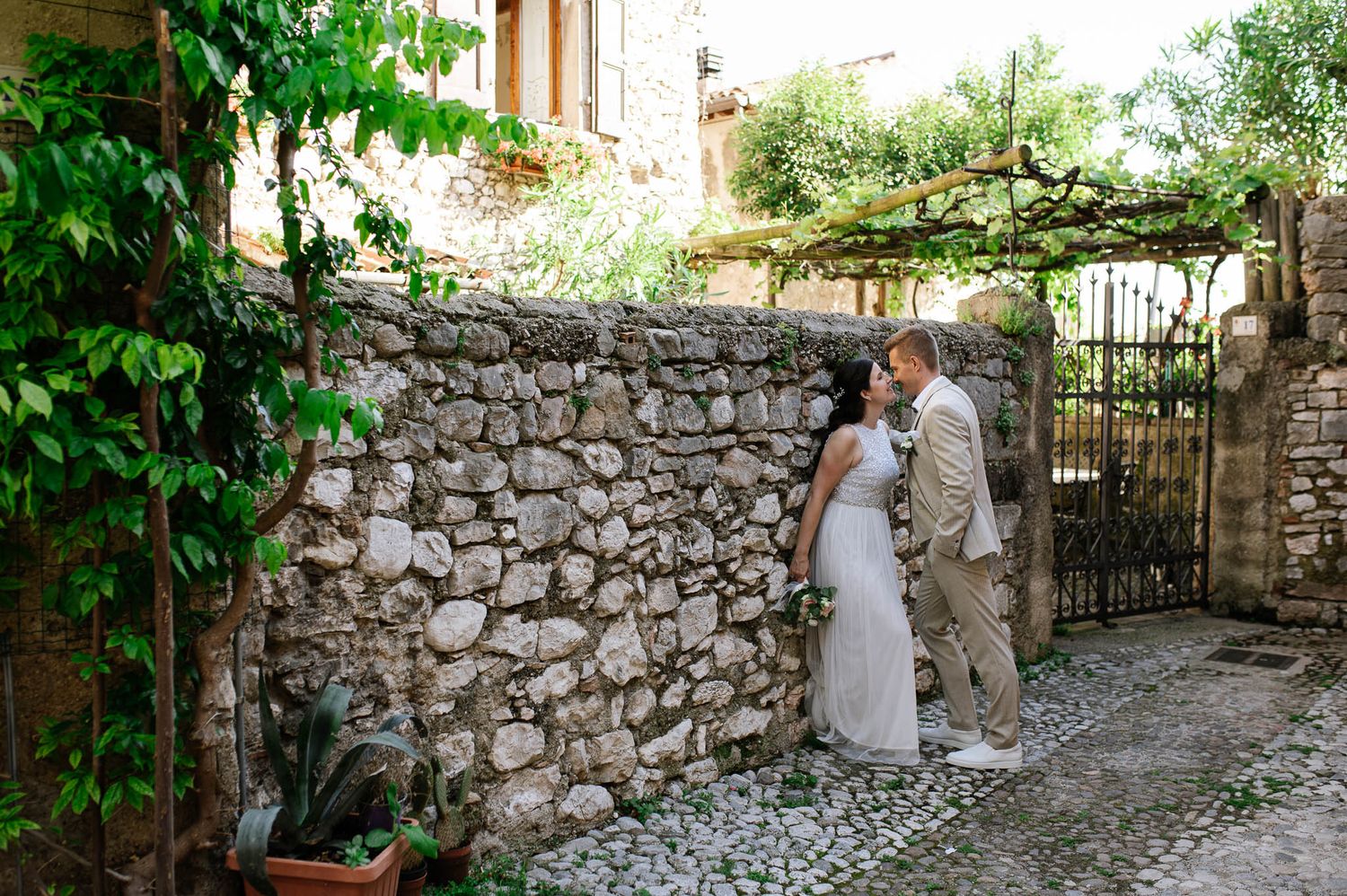 Newlyweds share a tender kiss beside rustic stone wall with iron gate in charming Italian village.