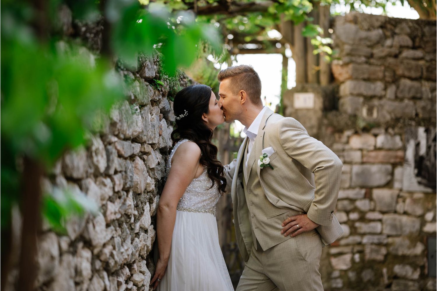 A bride and groom share a romantic kiss beside a rustic stone wall surrounded by lush green vines.