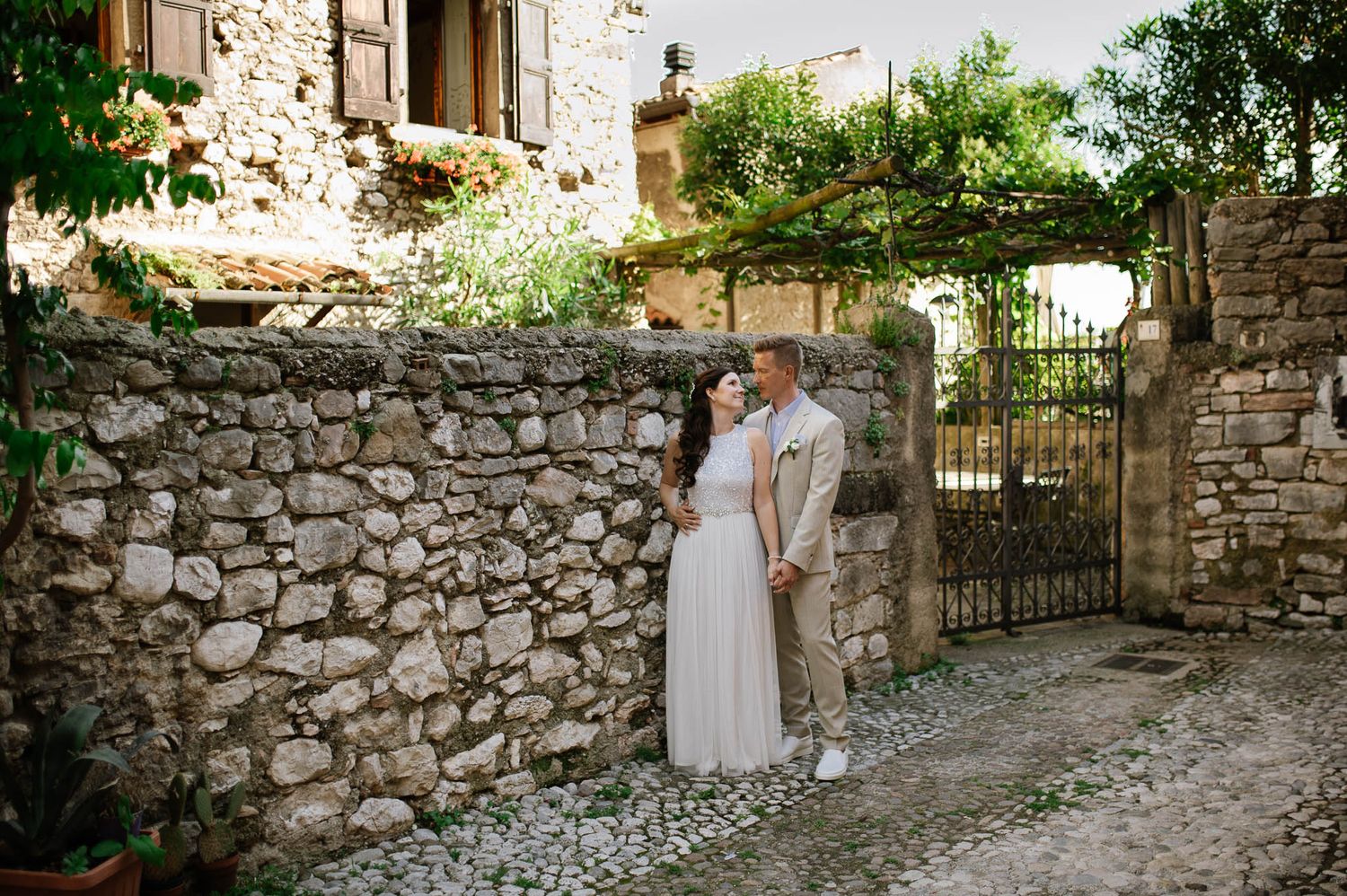 Bride and groom standing together by a rustic stone wall and iron gate in a charming village.