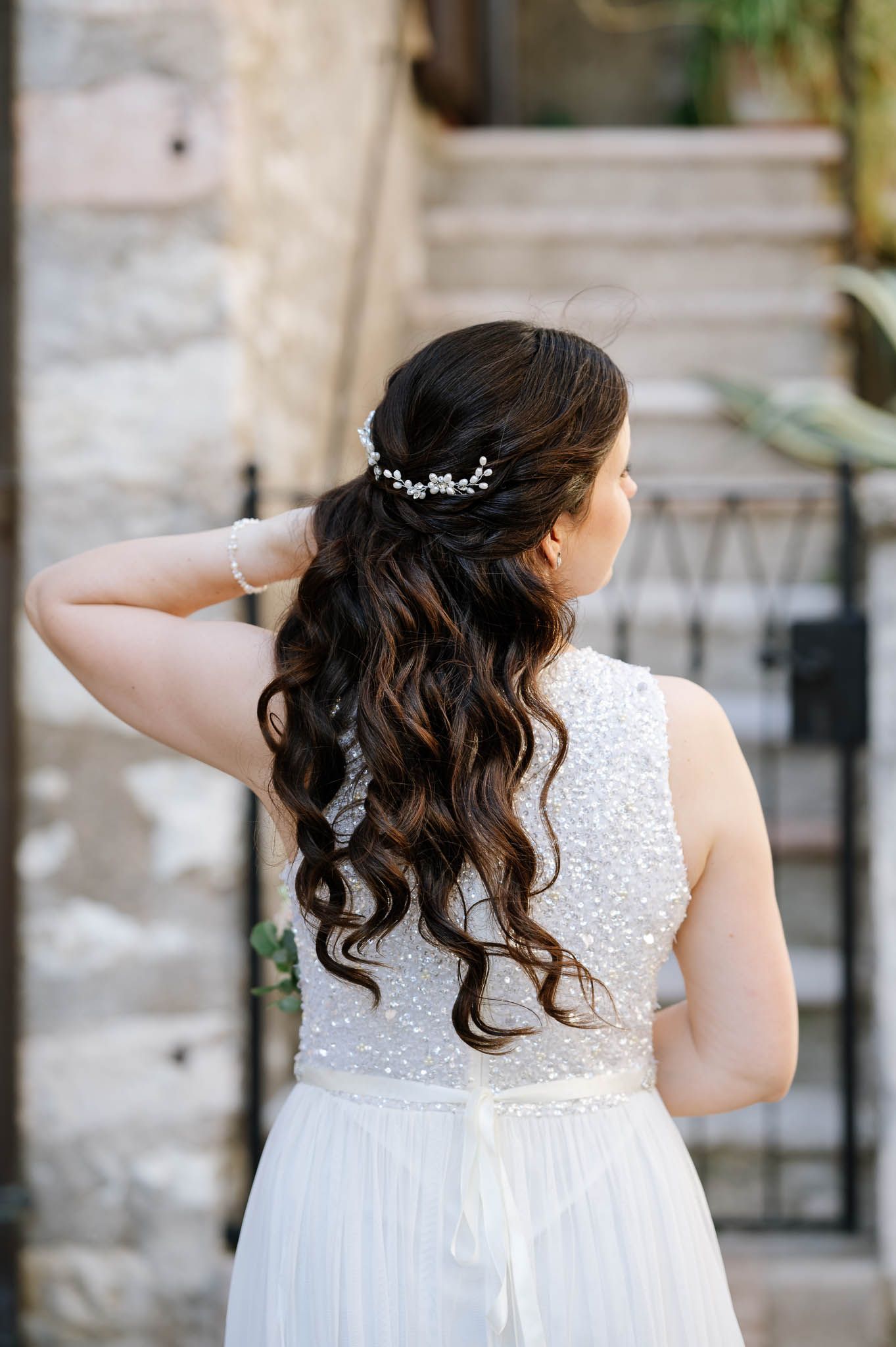 Back view of bride in beaded sleeveless gown with elegant half-up curly hairstyle adorned with pearl hair vine.