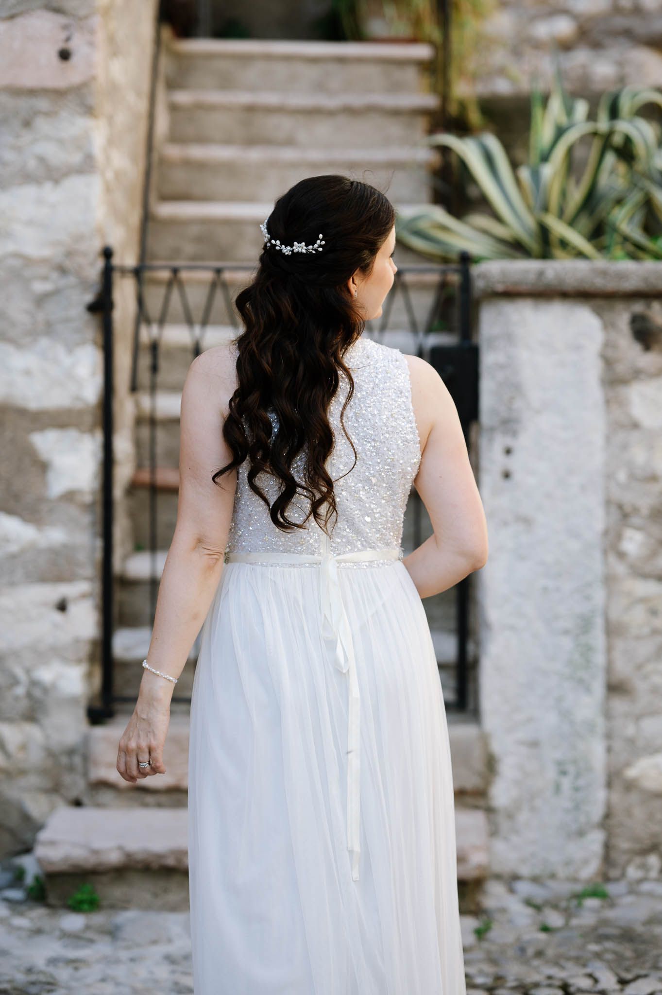 Bride in beaded white gown with long dark curly hair and floral hair accessory viewed from behind.