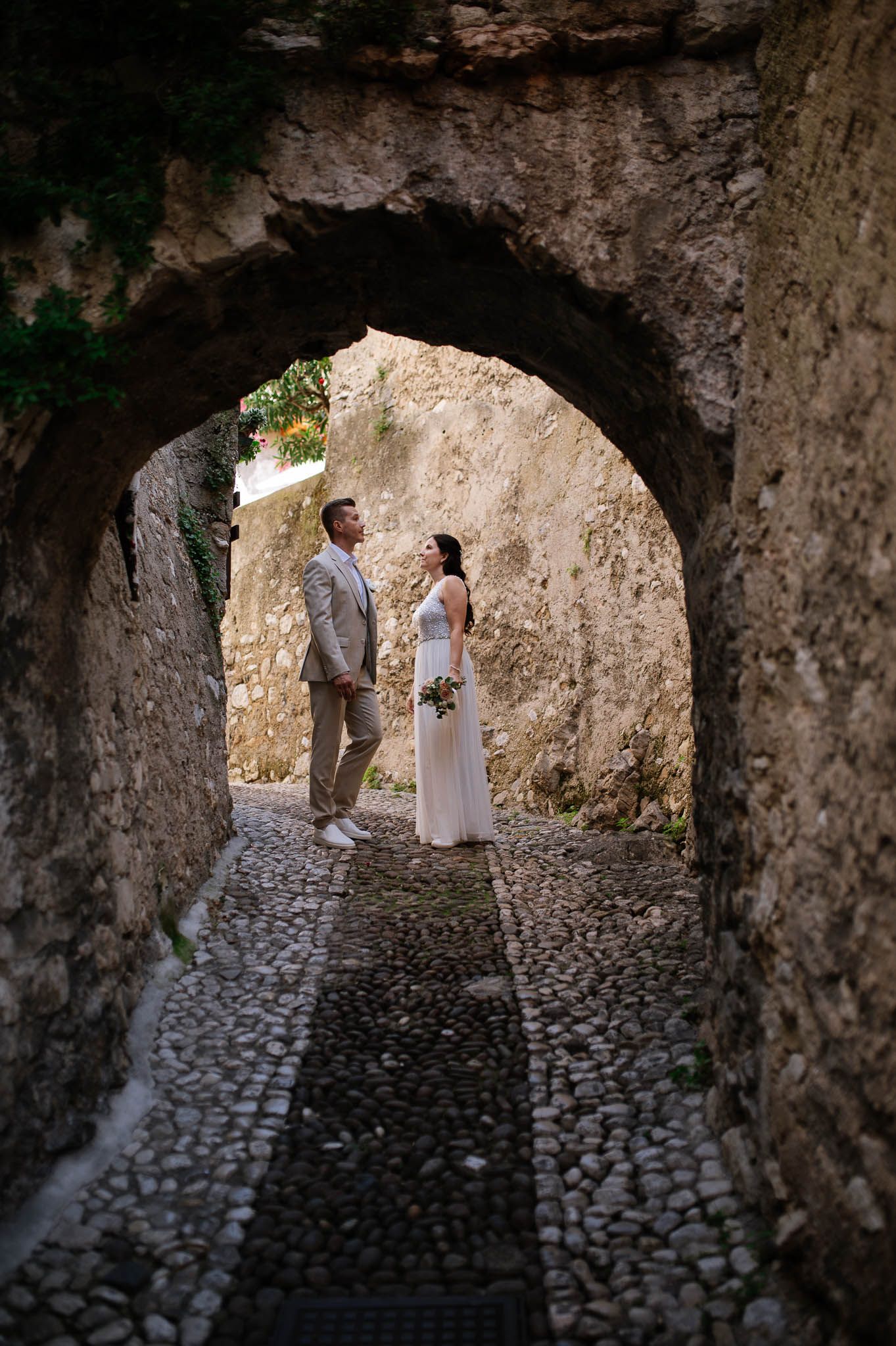 Bride and groom stand facing each other under a stone archway on cobblestone path in a historic European village.