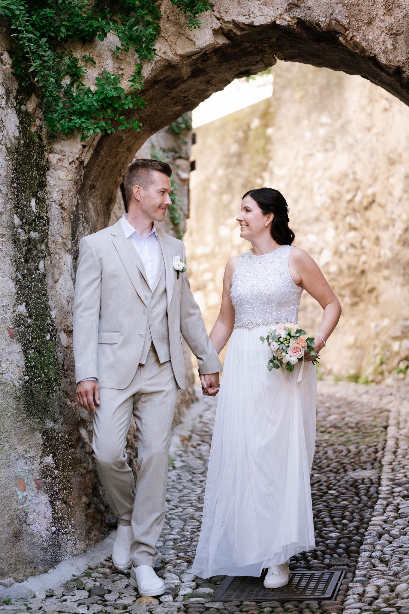 Smiling bride and groom walk hand in hand through a charming cobblestone archway on their wedding day.