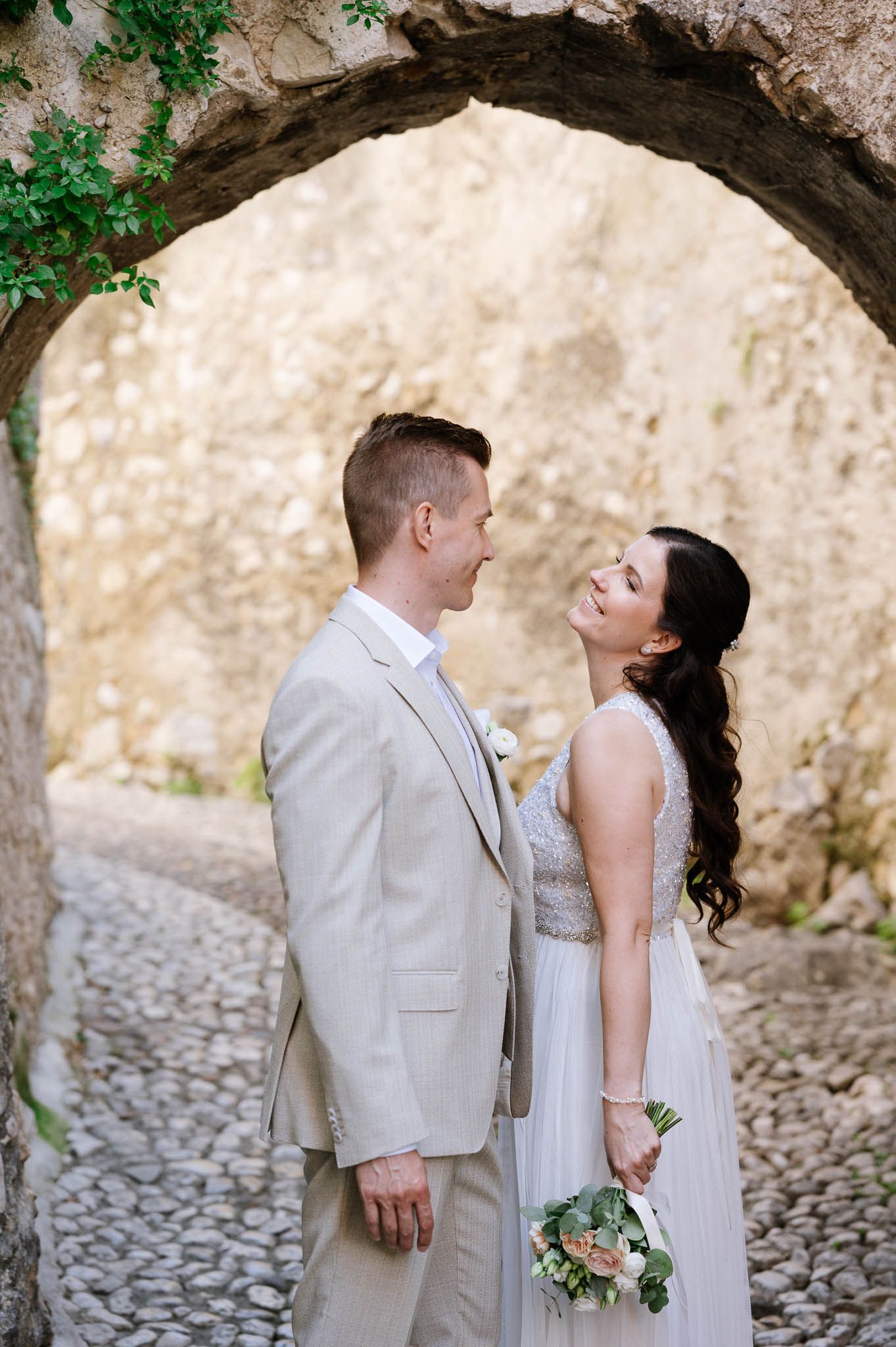 Laughing bride and groom stand under a rustic stone arch on cobblestone path at wedding.