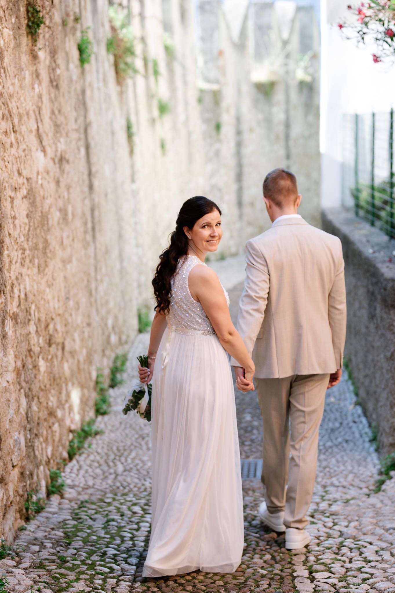 Bride in white gown looks back smiling while holding bouquet as couple walks cobblestone alley.
