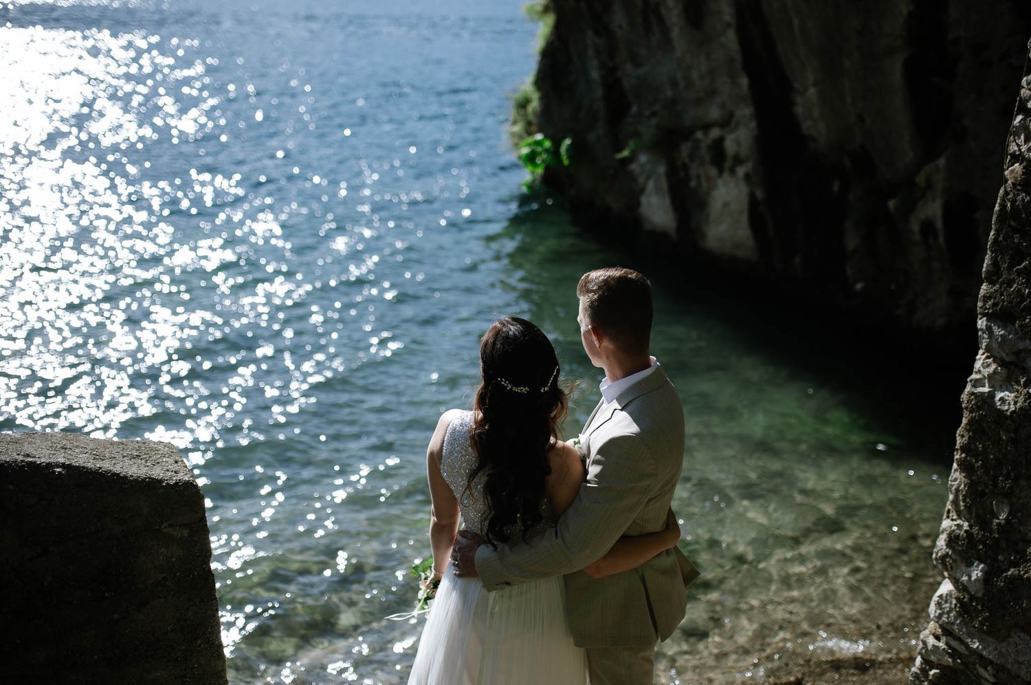 Newlywed couple embracing by sparkling turquoise lake waters beside ancient stone cliffs on their wedding day.