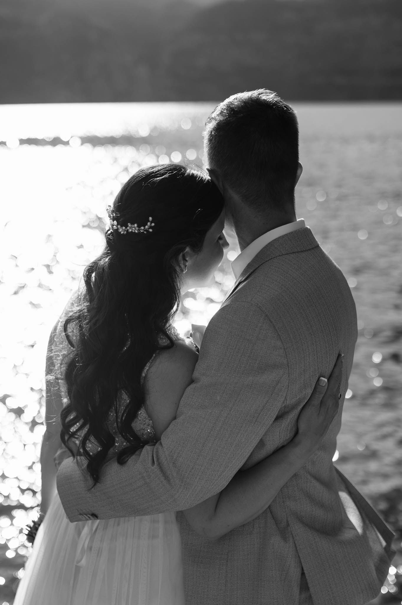 Black and white silhouette of bride and groom embracing romantically with sparkling sunlit lake in the background.