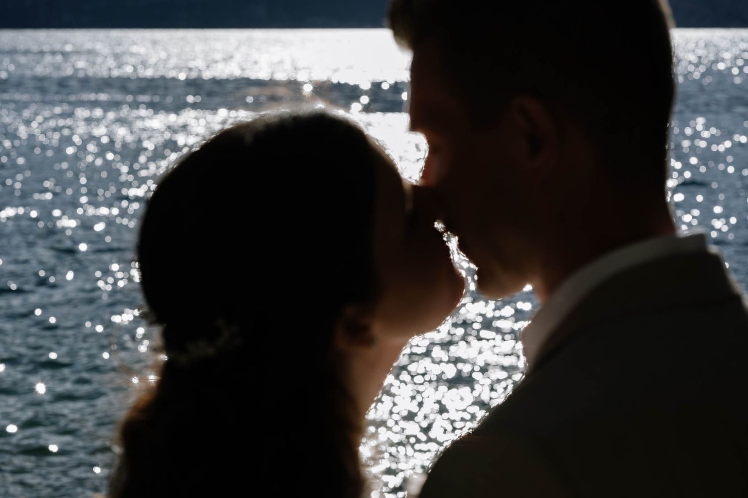 Romantic silhouette of bride and groom kissing against sparkling sunlit lake at dusk.