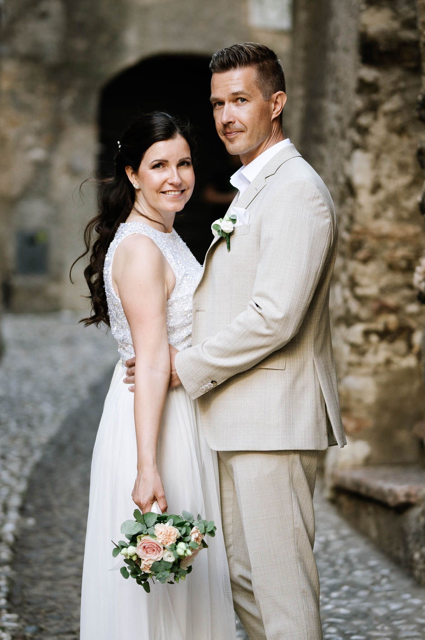 Smiling bride holding bouquet and groom in beige suit pose together in historic stone courtyard.