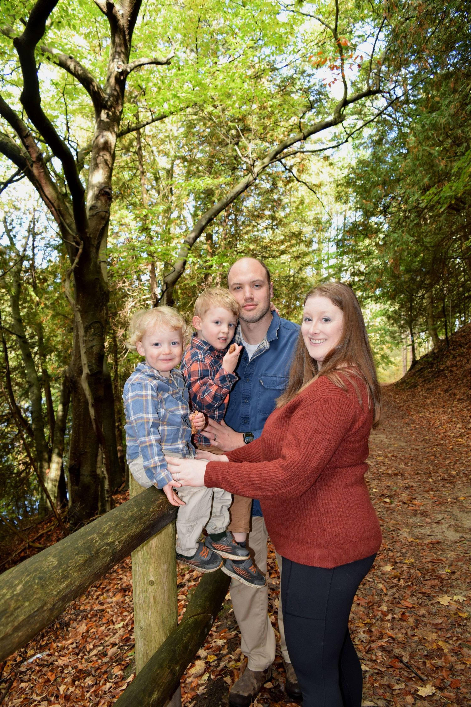 Family of four posing on a wooden fence trail surrounded by autumn foliage in a lush forest setting.
