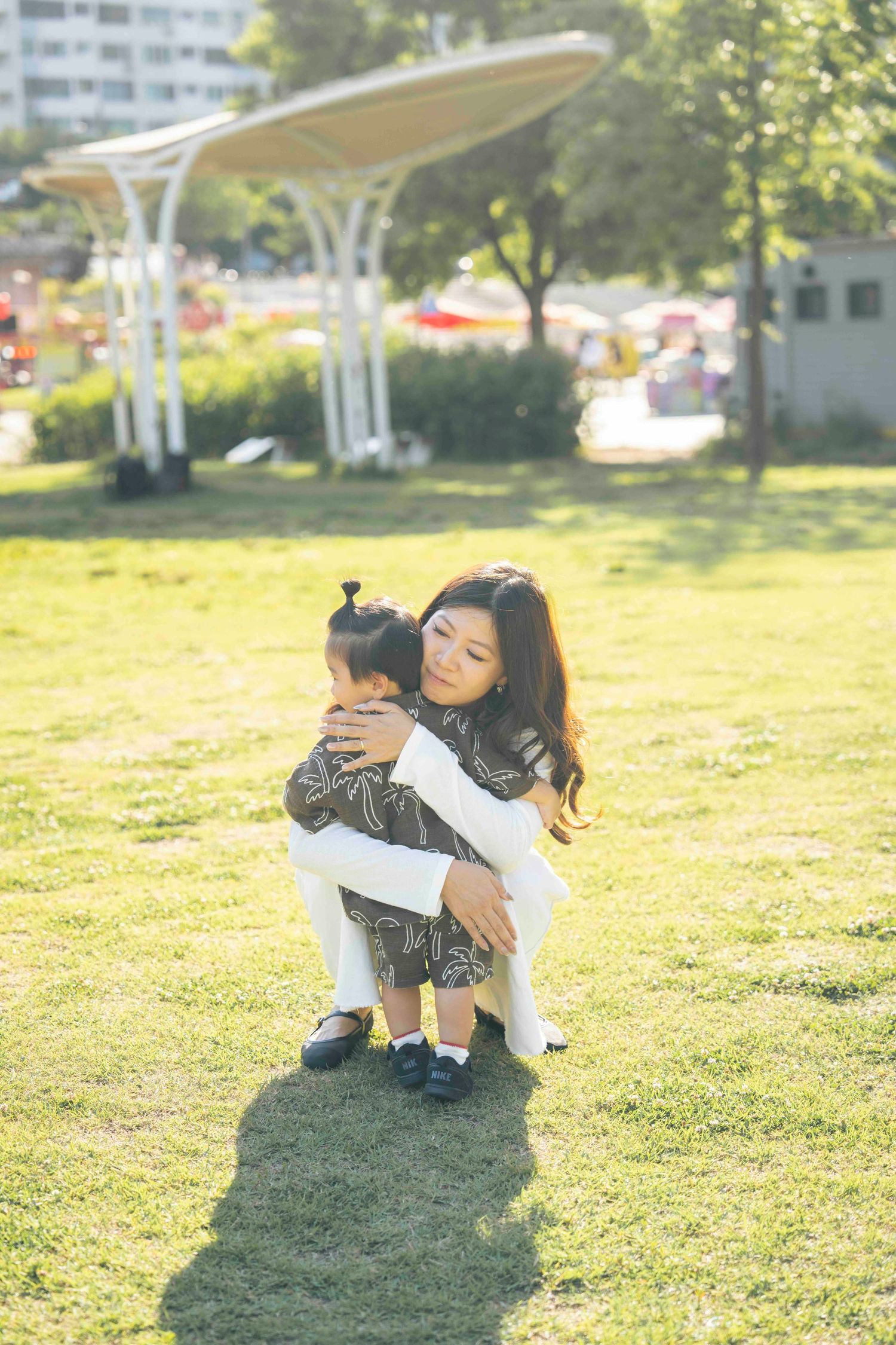A mother tenderly hugs her young child in a sunny green park, both dressed in stylish outfits.