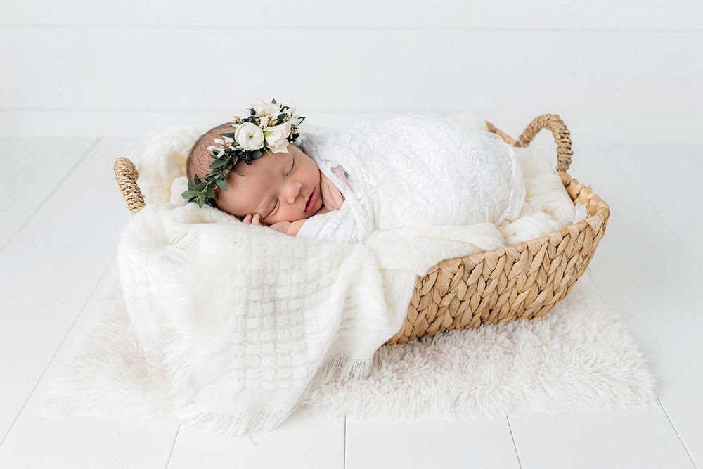 Sleeping newborn baby wrapped in white swaddle with floral crown in wicker basket on fluffy white rug.