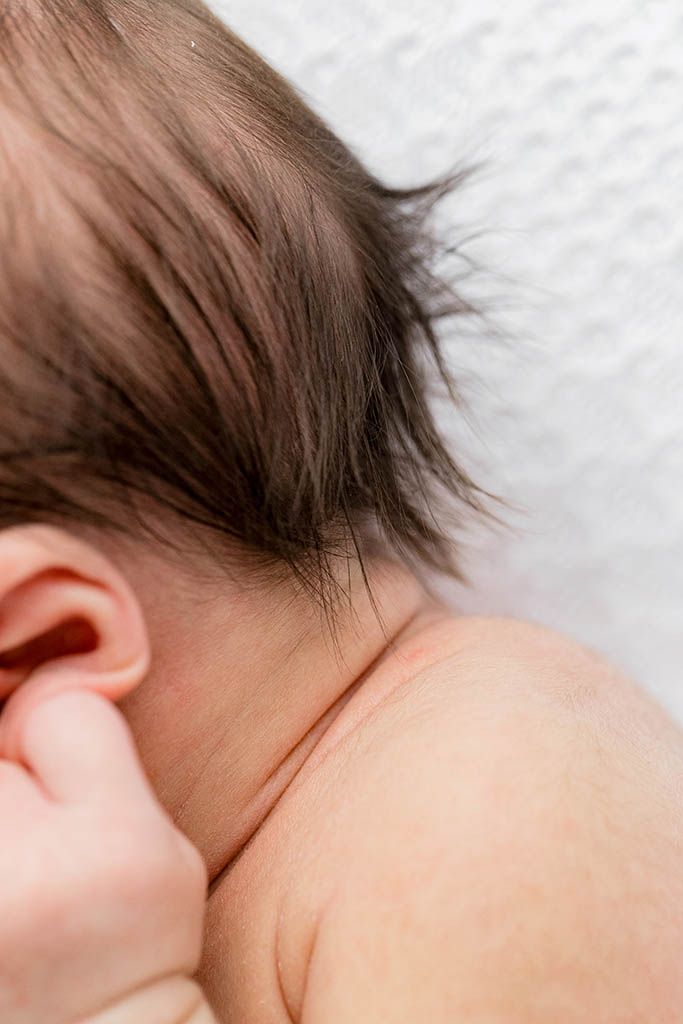 Close-up of a newborn baby's neck and ear with fine dark hair, resting peacefully against a white background.