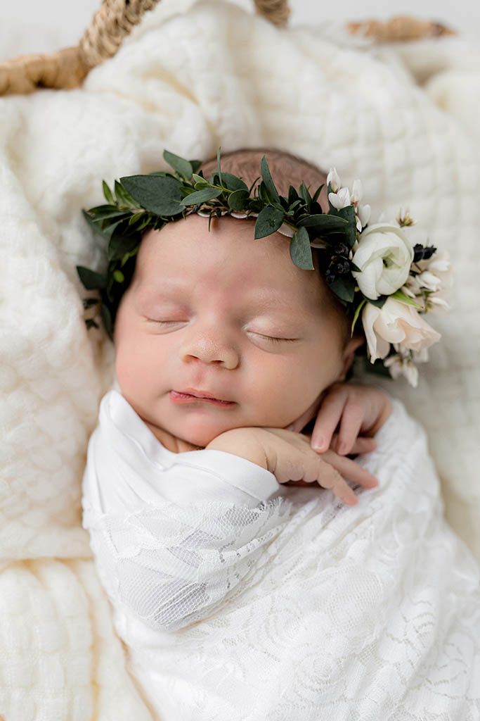 Sleeping newborn baby wrapped in white swaddle wearing a delicate green and white floral crown in a basket.
