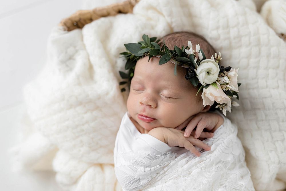 Sleeping newborn baby wrapped in white swaddle wearing a delicate floral crown in a cozy basket.