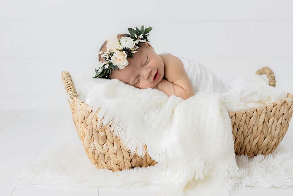 Sleeping newborn baby girl in floral crown resting in wicker basket with white fur blanket.