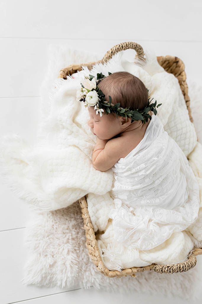 Newborn baby sleeping in a wicker basket with white blankets, wearing a delicate floral crown with white roses and greenery.