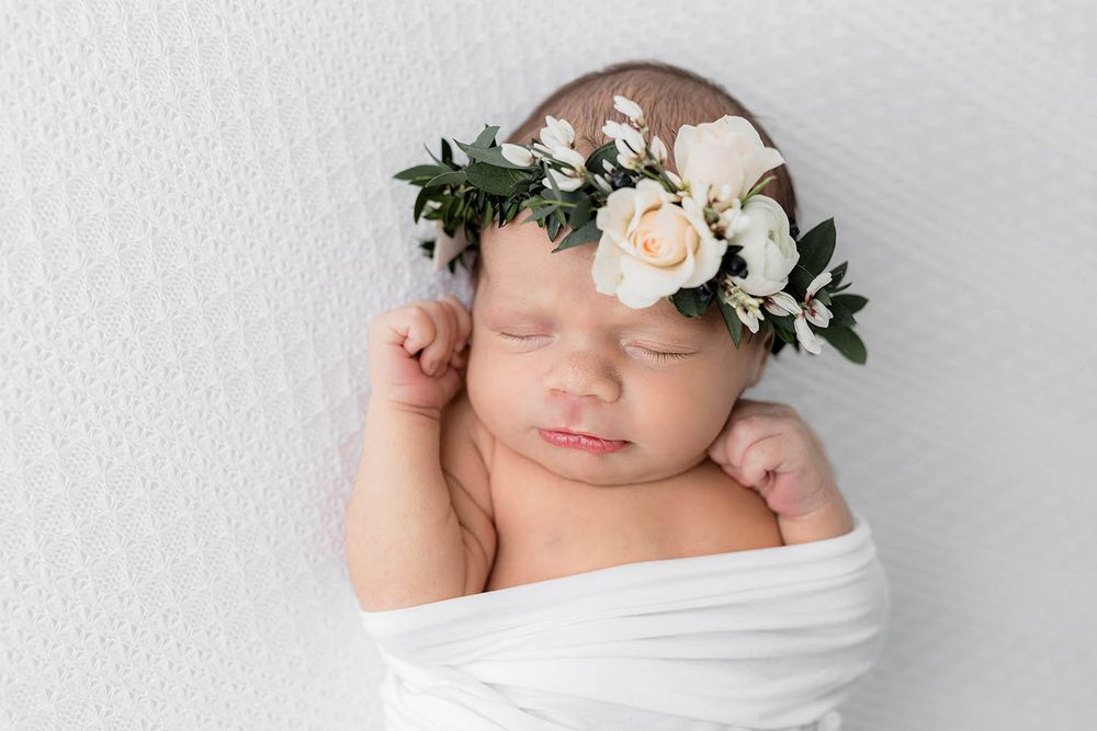 Peaceful newborn baby swaddled in white wrap wearing a delicate floral crown with roses and greenery.