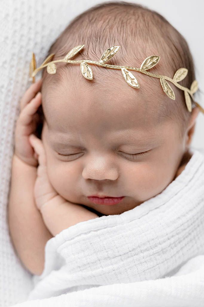 Sleeping newborn baby wrapped in white swaddle wearing a delicate gold leaf crown headband.