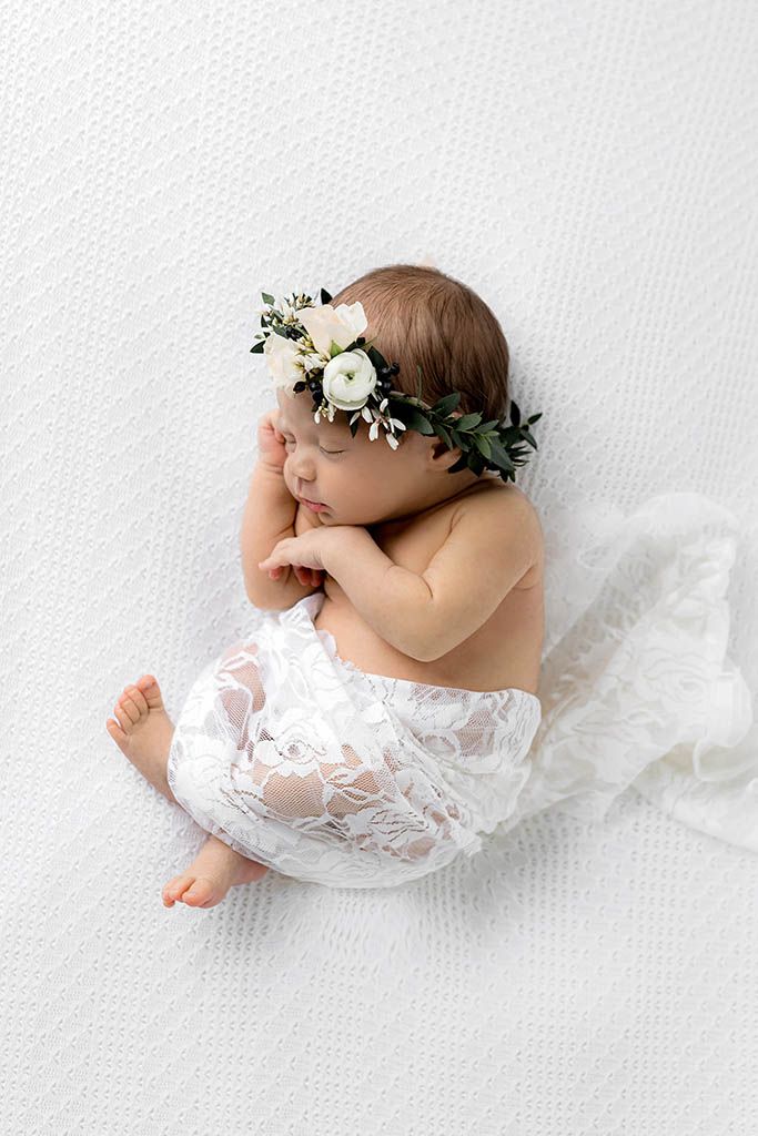 Sleeping newborn baby wearing a white floral crown and lace wrap, posed on a soft white blanket.