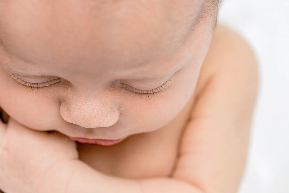 Close-up of a sleeping newborn baby with tiny eyelashes resting chin on hand against white background.