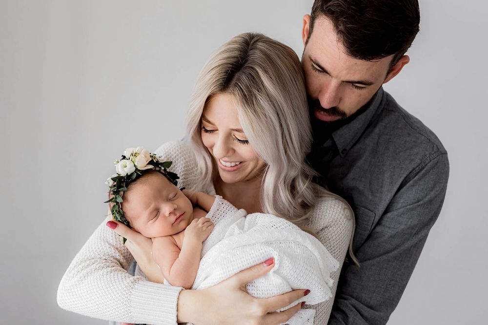 New parents lovingly cradle their sleeping newborn baby girl wearing a floral crown in a studio portrait.