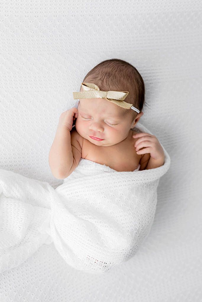 Peaceful newborn baby swaddled in white wrap with a small gold bow headband sleeping on white backdrop.