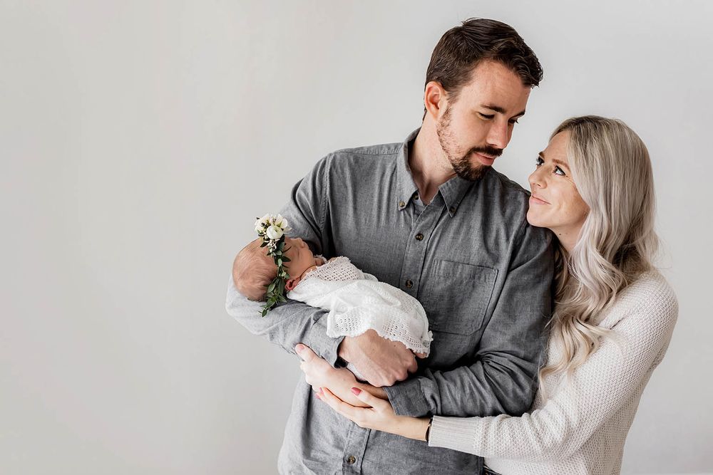 Happy couple smiling at their newborn baby with a floral headband in a bright studio photo.