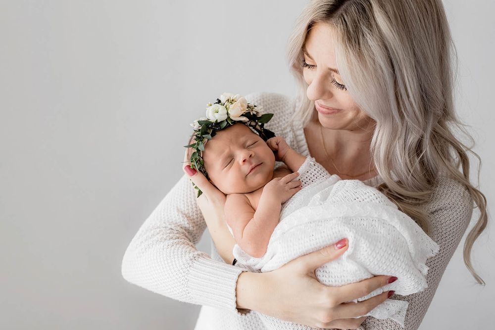 Loving mother in white sweater gently holding sleeping newborn baby wearing a beautiful floral crown.