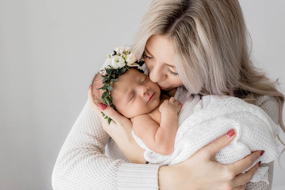 Mother tenderly kissing her sleeping newborn wearing a delicate floral crown wrapped in white.
