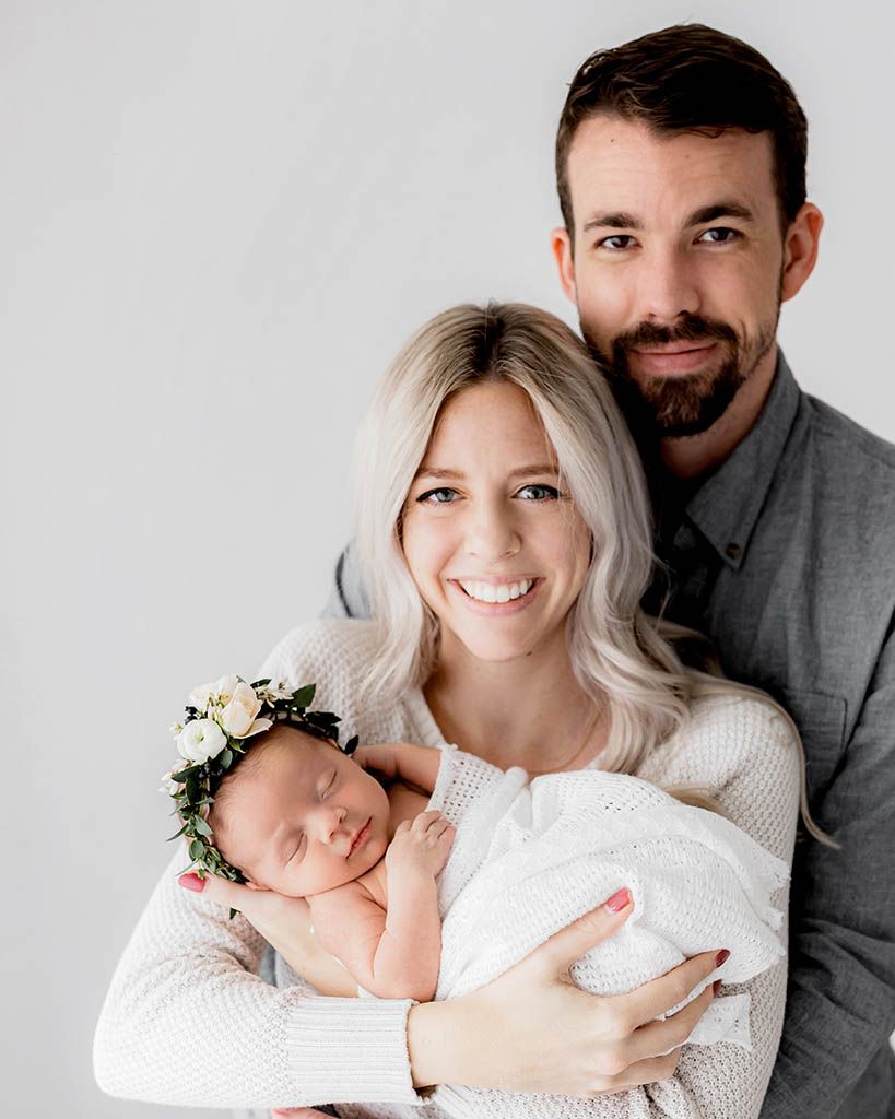 Happy parents smiling and holding their newborn baby girl wearing a white floral crown.