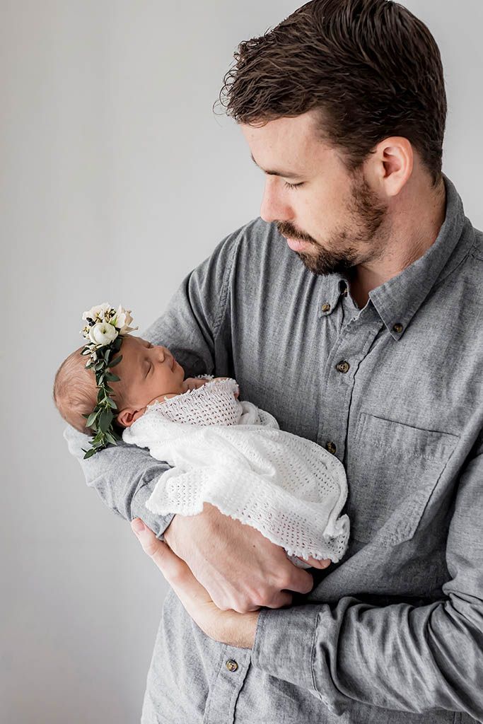 Proud father in gray shirt tenderly holding his sleeping newborn baby girl in floral crown.