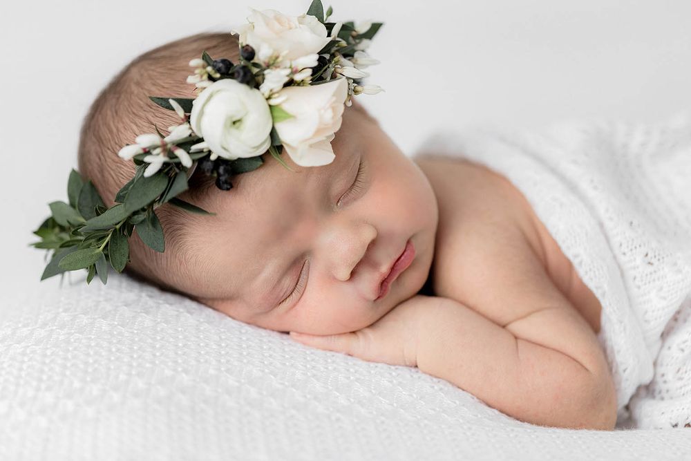 Peaceful sleeping newborn baby girl wearing a white floral crown on a white blanket.