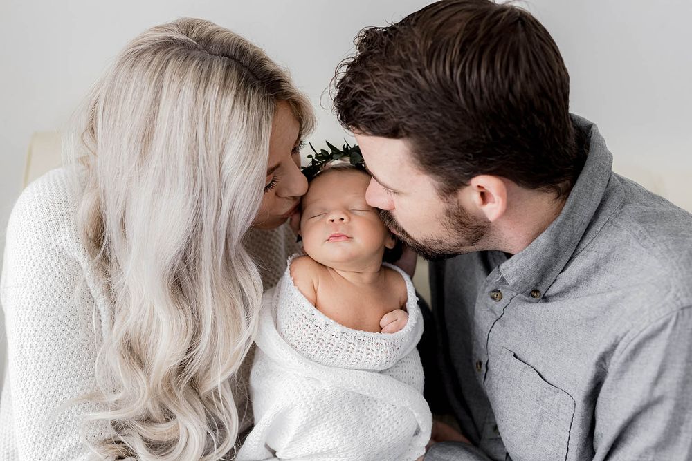 New parents tenderly kiss their swaddled newborn baby on the forehead in a white studio setting.