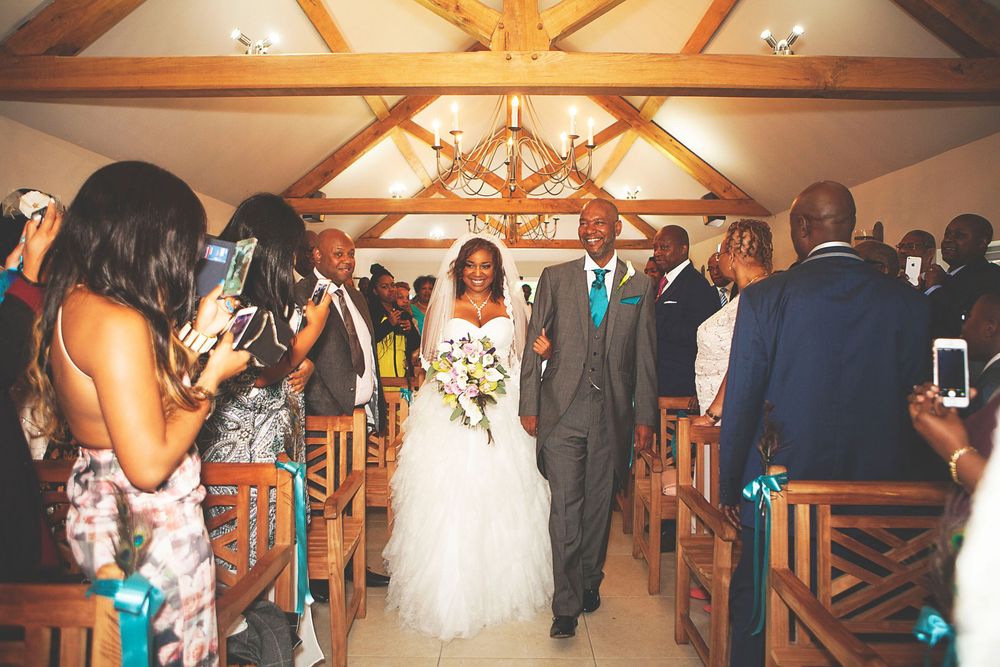 Bride and groom walking down the aisle in a rustic chapel, surrounded by guests capturing the wedding ceremony. At Oaks farm wedding venue.
